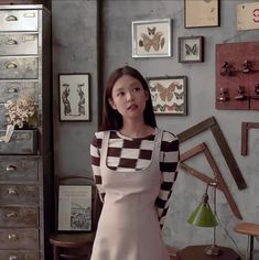 a woman standing in front of a dresser with drawers and pictures on the wall behind her