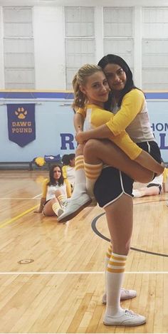 two girls in cheer uniforms hugging each other on a basketball court while others watch from the sidelines