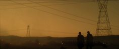 two men standing in the middle of an empty field at sunset with power lines in the background