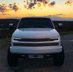 the front end of a white truck parked in a parking lot at sunset or dawn