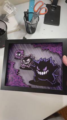 a person holding up a pixel art piece with scissors and other items in the background