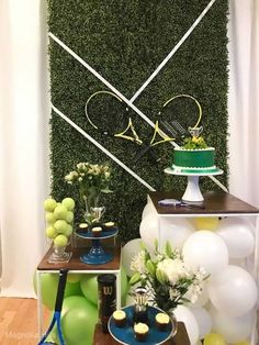 a table topped with cake and balloons next to a wall covered in greenery that has tennis rackets on it