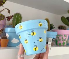 a hand holding up a blue flower pot with bees on it and cactus in the background