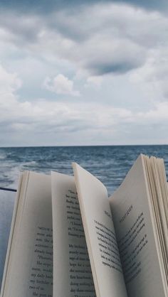 an open book sitting on top of a wooden table next to the ocean under a cloudy sky