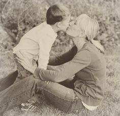 a woman kissing a young boy on the cheek while sitting in a field with tall grass