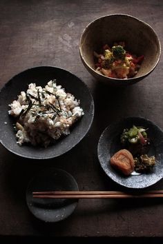 three black bowls filled with food next to chopsticks on top of a wooden table