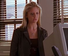 a woman standing in front of a computer monitor with blinds on the windows behind her