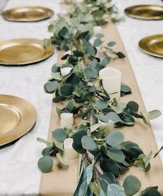 the table is set with gold plates and greenery on it, along with candles