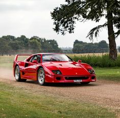 a red sports car driving down a dirt road next to a tree and grass covered field