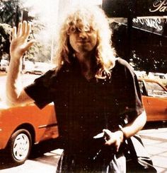 a man with long hair standing in front of a red car waving at the camera