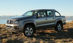 a silver truck parked on top of a dry grass field next to the ocean with mountains in the background