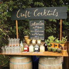 a wooden table topped with lots of bottles and glasses next to barrels filled with plants