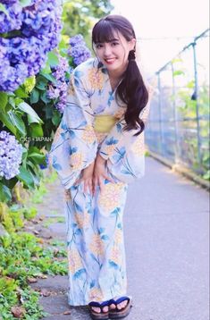 a woman standing on the side of a road next to purple flowers