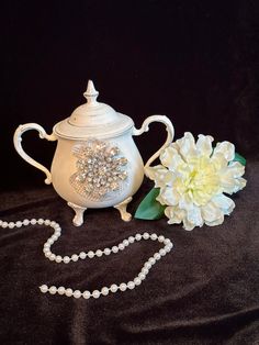 a white teapot with pearls and a flower on a black tablecloth next to it