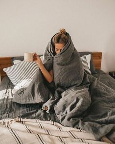 a woman laying in bed wrapped up with a blanket and holding a coffee cup while looking at her phone