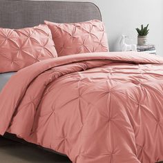 a bed with pink comforter and pillows in a room next to a plant on a table