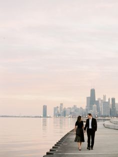 a man and woman are walking along the water in front of a cityscape