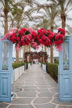 an archway decorated with pink flowers and greenery on the side of a walkway between two palm trees