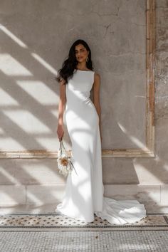 a woman standing in front of a wall wearing a white dress and holding a bouquet