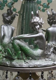 two little mermaid figurines sitting on top of a silver plate with green beads