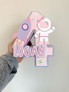 a person holding up a pink and purple number four shaped like a rocket ship with the word logan on it