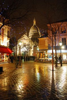 people are walking down the street at night in front of buildings with lights on them