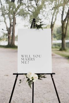 a sign that says will you marry me? with flowers on it in front of some trees