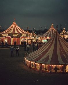 a large circus tent with lights on it's sides and people walking around the tents