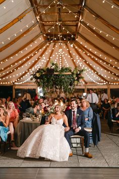 a bride and groom sitting at their wedding reception in a marquee with string lights