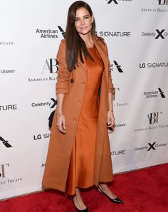 a woman standing on a red carpet wearing an orange dress and trench coat with black heels