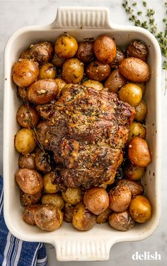 meat and potatoes in a white casserole dish on a blue towel next to some herbs