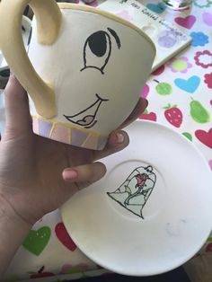 a hand holding a coffee cup with a face drawn on the side and a plate next to it