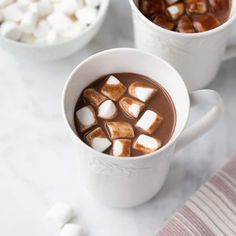 two mugs filled with hot chocolate and marshmallows on a white table