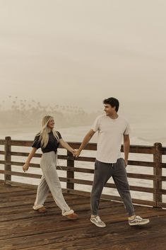 a man and woman holding hands while walking on a boardwalk near the ocean with fog in the background