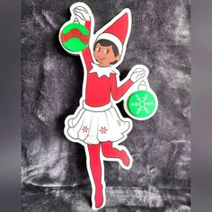 the elf is holding a green ornament in one hand and wearing a red hat