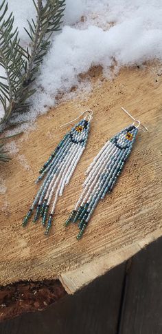 two beaded earrings sitting on top of a piece of wood next to a pine tree