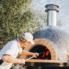 a man in white shirt and cap cooking food over an outdoor brick oven with fire