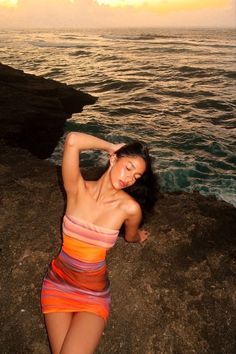 a beautiful woman in a colorful dress standing on the rocks by the ocean at sunset