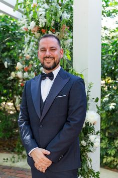 a man in a tuxedo smiles at the camera while standing under a floral arch