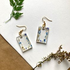 two square glass earrings sitting on top of a piece of paper next to a plant