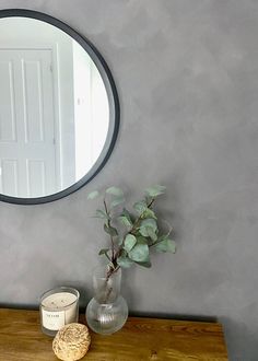 a round mirror on the wall above a wooden table with a vase and candle next to it