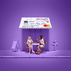 two women sitting at a table in front of a purple wall with an advertisement on it