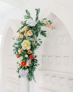 an arrangement of flowers and greenery is displayed on a white pedestal in front of wine glasses