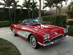 an old red convertible car parked in front of a palm tree lined driveway with bushes and trees