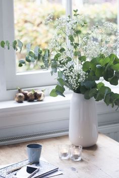 a white vase filled with lots of flowers on top of a table next to a window