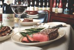 two plates with steak, asparagus and potatoes next to wine bottles on a table