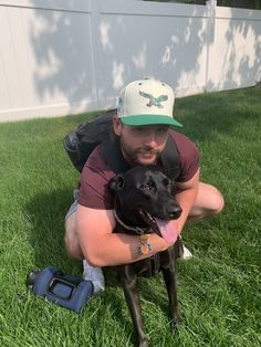 a man kneeling in the grass holding a black dog with his tongue out and wearing a hat