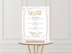 a pink and gold wedding menu card on a wooden table with a white wall in the background