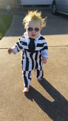 a toddler dressed up in a costume and tie walking down the street with his shadow on the ground