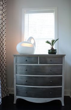 a white vase sitting on top of a dresser next to a window with curtains behind it